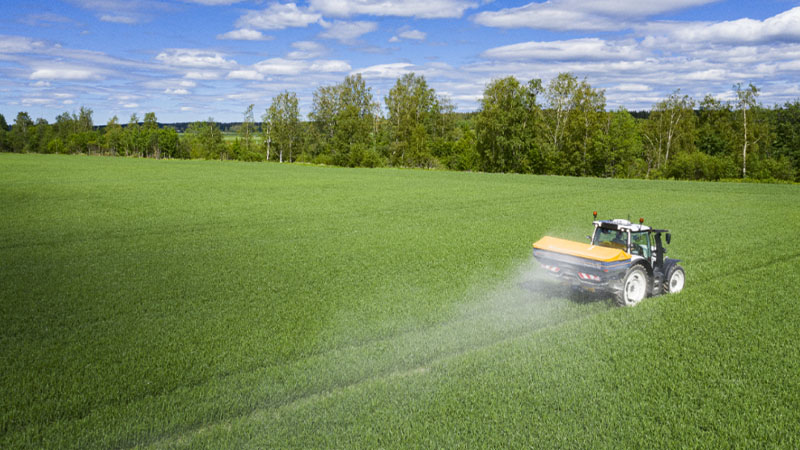 Valtra tractor is operating in a large green field while carrying out fertilizer spreading. The image shows the tractor moving across a growing crop, with the spread pattern visible behind the machine. This application supports efficient nutrient distribution and crop management during the growing season. The open field environment highlights the tractor's role in productive arable farming operations and field efficiency.