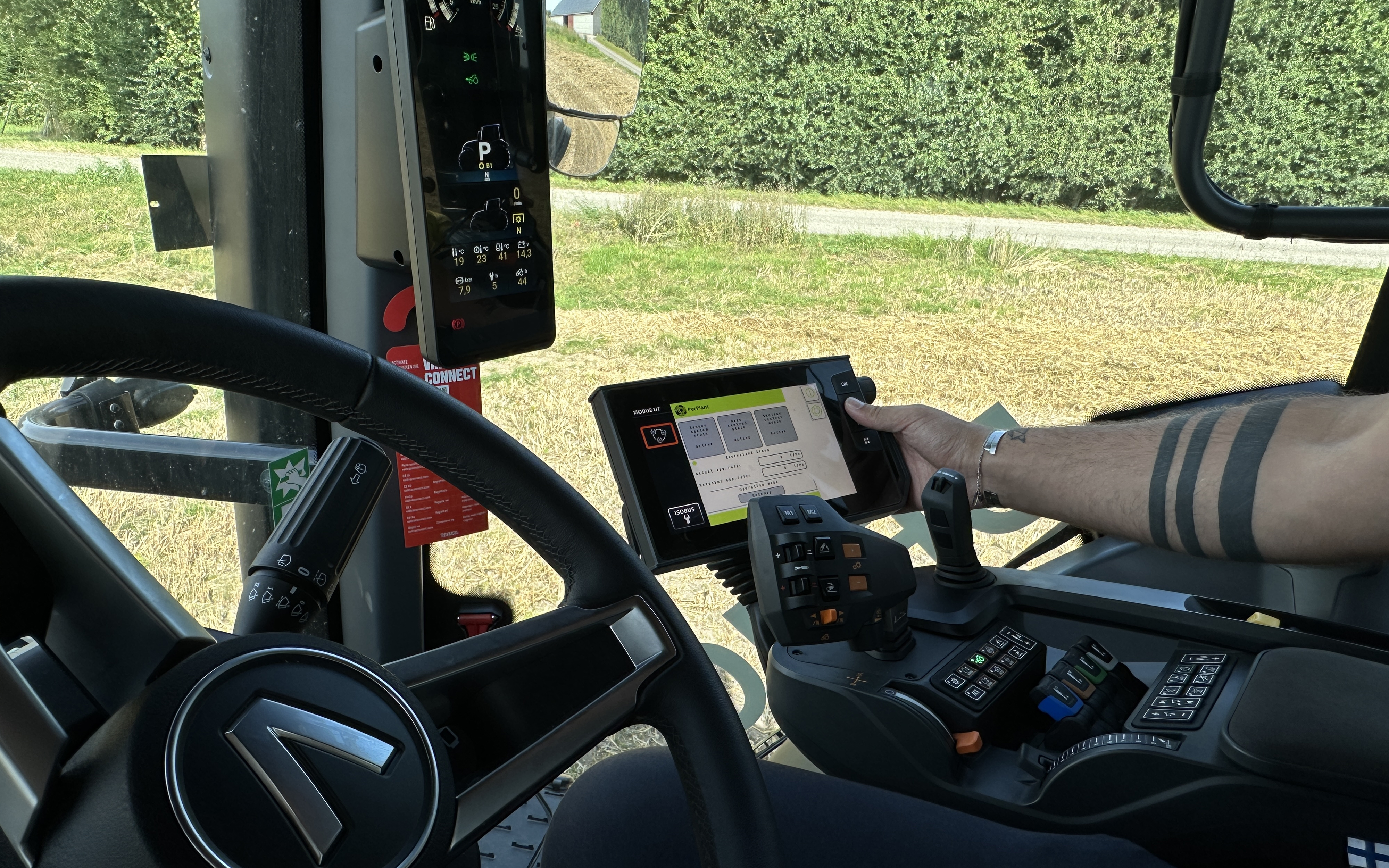 The interior cab of a Valtra Q Series  tractor with a driver’s arm operating a SmartTouch touchscreen control panel. Multiple digital displays, buttons, and joysticks are visible, indicating advanced agricultural machinery controls. Outside the cab, a rural field and hedges can be seen through the windows. 