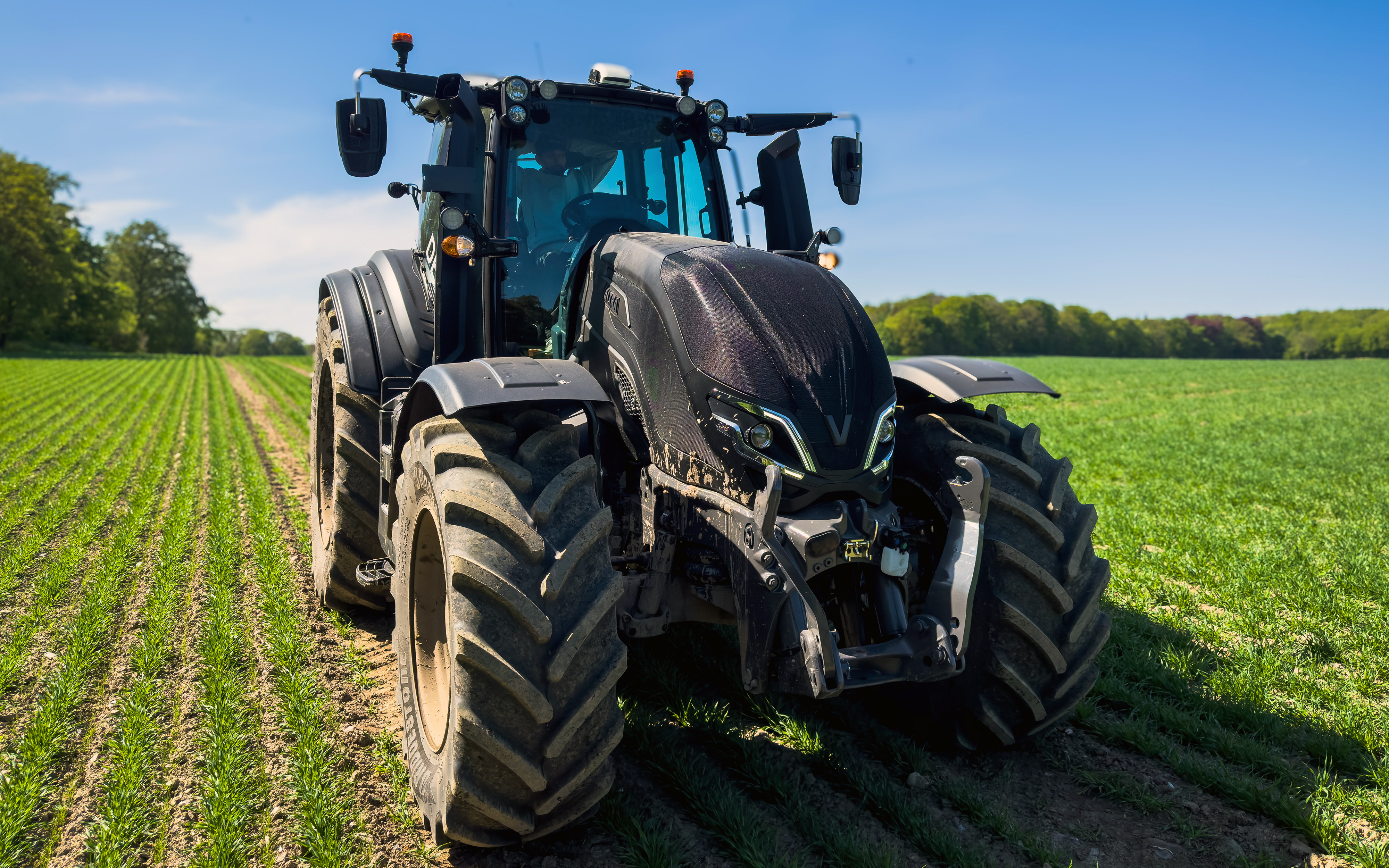 A large black Valtra Q Series tractor is positioned in the middle of a cultivated field with young green crops. The vehicle is shown from the front, highlighting its oversized tires and robust machinery. 