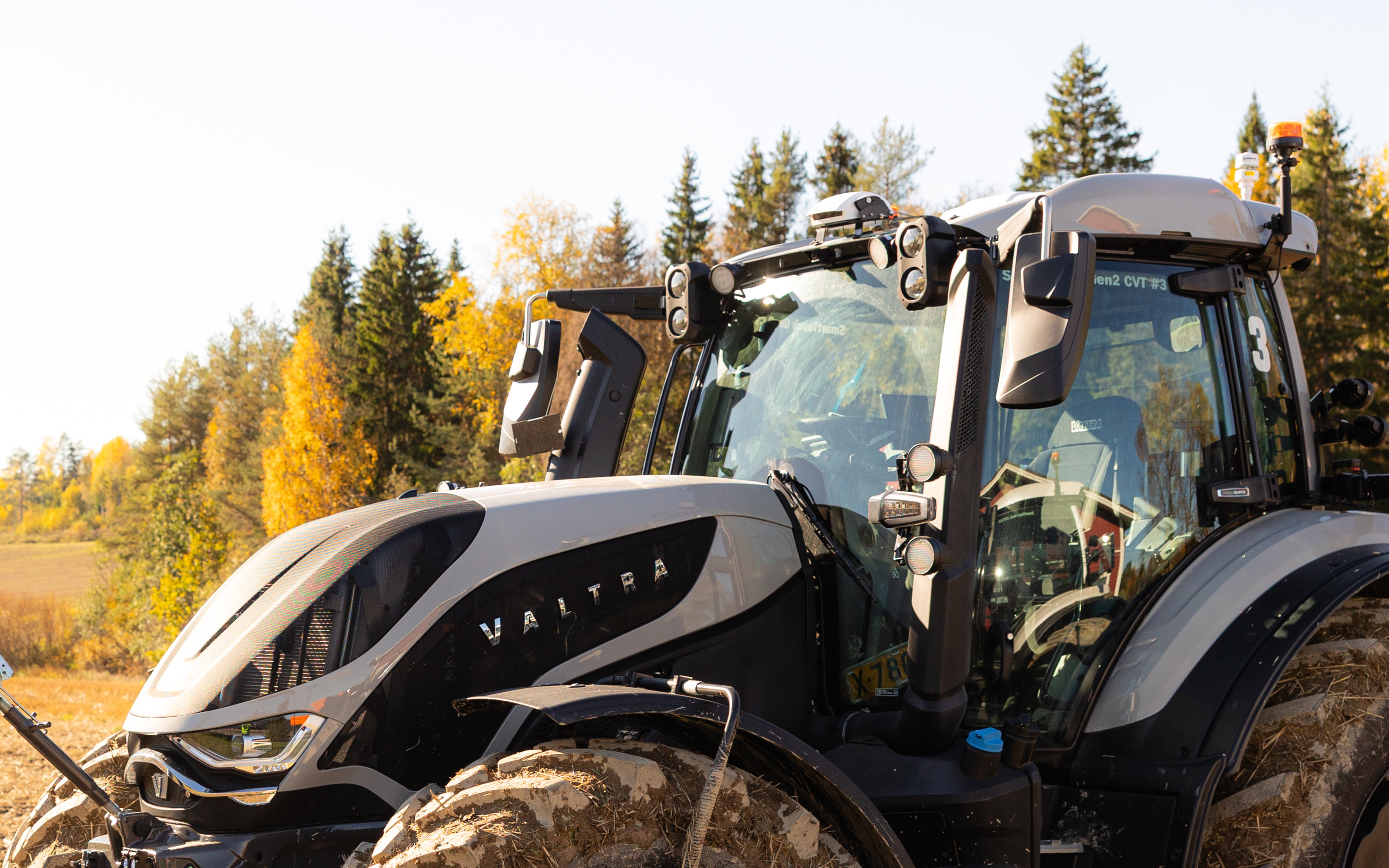 A Valtra T Series tractor is shown from a close side angle in a rural field. On the roof of the cab a sensor box can be seen. The vehicle’s cab and front section are prominently visible, covered with mud from recent work. In the background, autumn-colored trees and open farmland suggest an agricultural setting.