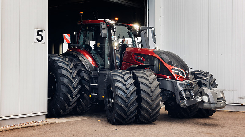 A modern, large red Valtra Q Series tractor with dual front and rear tires is seen emerging from an industrial building. The building is marked with a visible number '5' near the entrance. The tractor features a robust design, glossy finish, and prominent headlights, suggesting advanced agricultural machinery. The setting is industrial, with neutral tones and a focus on the vehicle's impressive size and engineering.