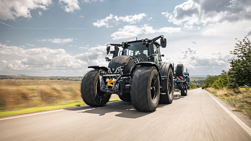 A large, modern black Valtra tractor is captured driving down a paved rural road. The vehicle dominates the foreground, with expansive countryside and cloudy skies in the background. The tractor's robust tires and mechanical details are clearly visible, emphasizing its power and utility. No visible text, numbers, or identifiable brands are present in the image.