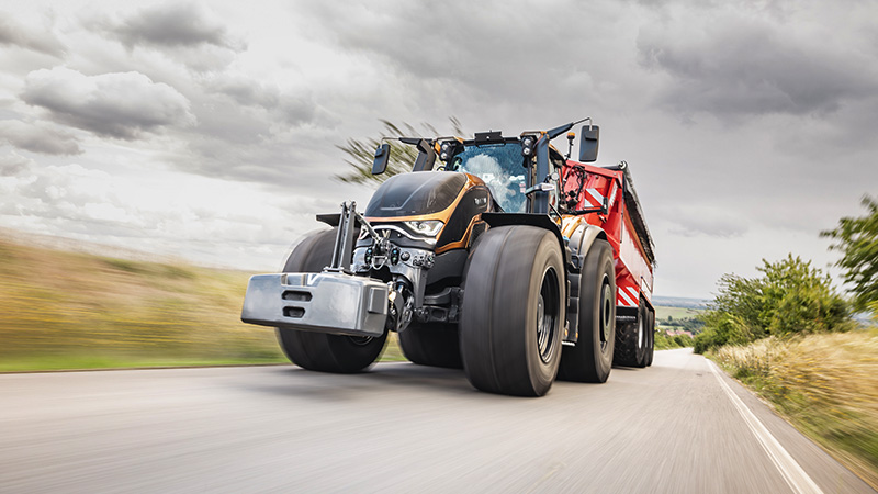 A large, modern black Valtra tractor is captured in motion, hauling a red trailer along a paved rural road. The scene is set under a dramatic, cloudy sky, with blurred grass and fields indicating speed. The tractor's robust design and oversized tires are prominent, emphasizing power and utility. No visible text, numbers, or identifiable brands are present in the image.
