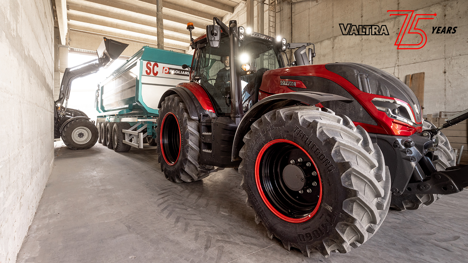 A large red and black Valtra tractor is parked inside an industrial warehouse, attached to a heavy-duty trailer. The vehicle is positioned close to a concrete wall, emphasizing its massive tires and robust construction. Bright daylight filters through the ceiling openings, highlighting the machinery and the teal and white trailer. The Valtra 75 years graphic appears in the upper right corner, marking an anniversary celebration.
